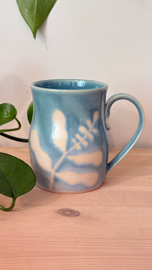 A light blue ceramic mug with a white painted leaf design, sitting on a light wood surface next to green plant leaves against a plain white wall.
